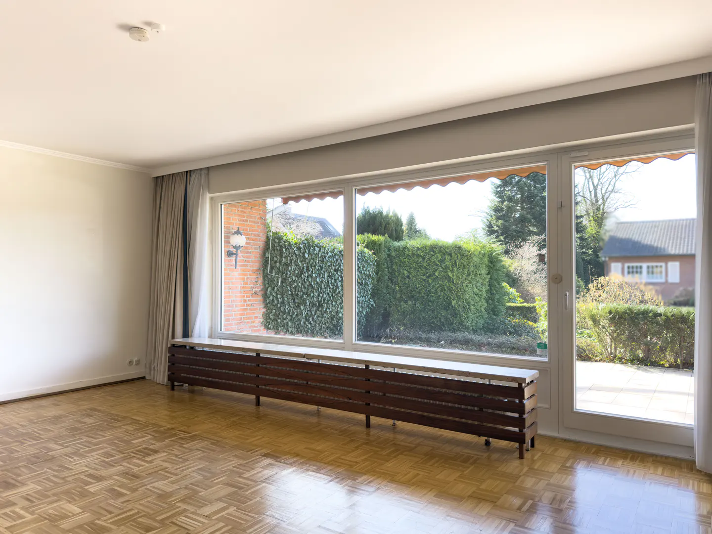 Bright, empty room with parquet floors, large windows overlooking a green garden, and a long, dark wood radiator cover.