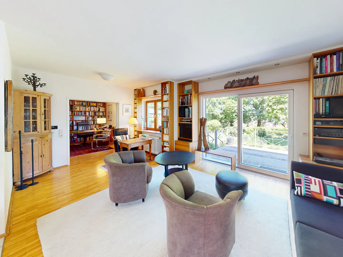 Bright living room with hardwood floors, white rug, brown armchairs, and a black coffee table. Bookshelves line the walls, and a sliding glass door leads to a patio.