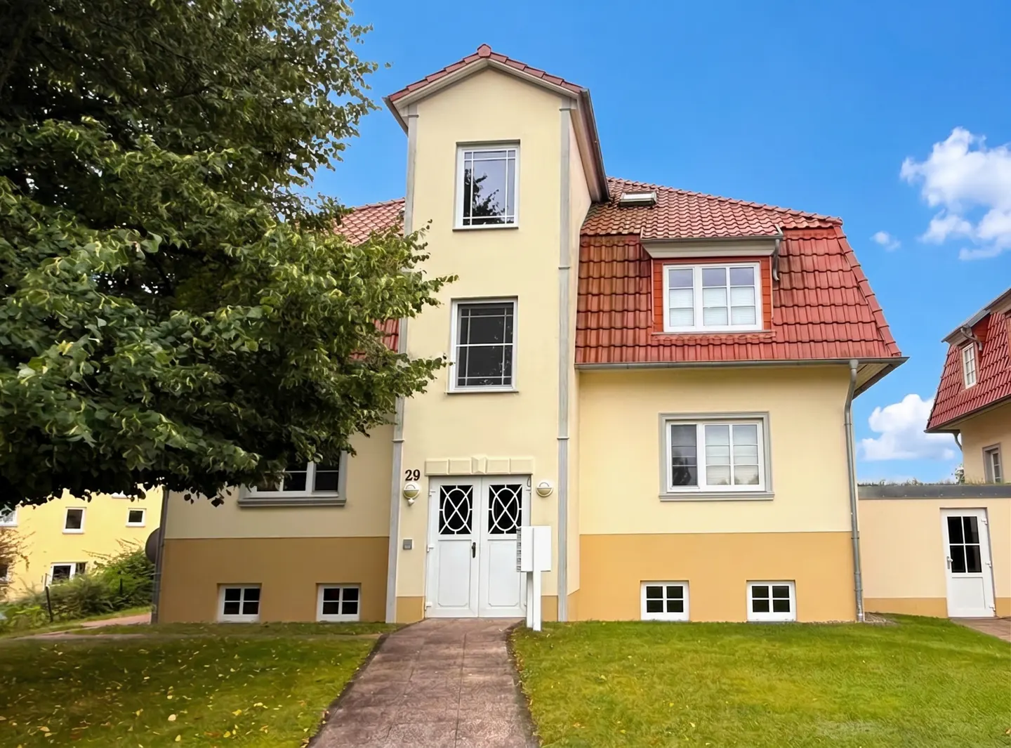 Two-story yellow house with a red tile roof, white trim, and white double doors at number 29. A tree is on the left.