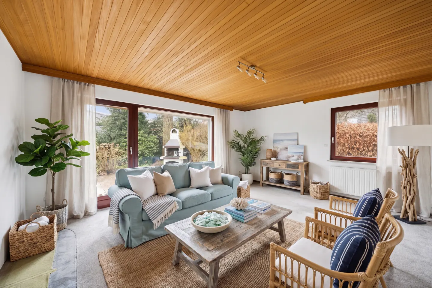 Living room with wood ceiling, light blue sofa, and rattan chairs. Large windows overlook a green yard with a grill. Coastal decor accents the space.