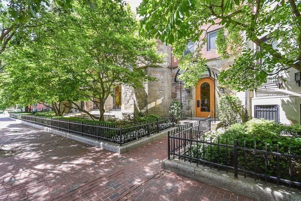 Exterior view of a stone building with a wooden door, green trees, and a brick walkway with a black metal fence.