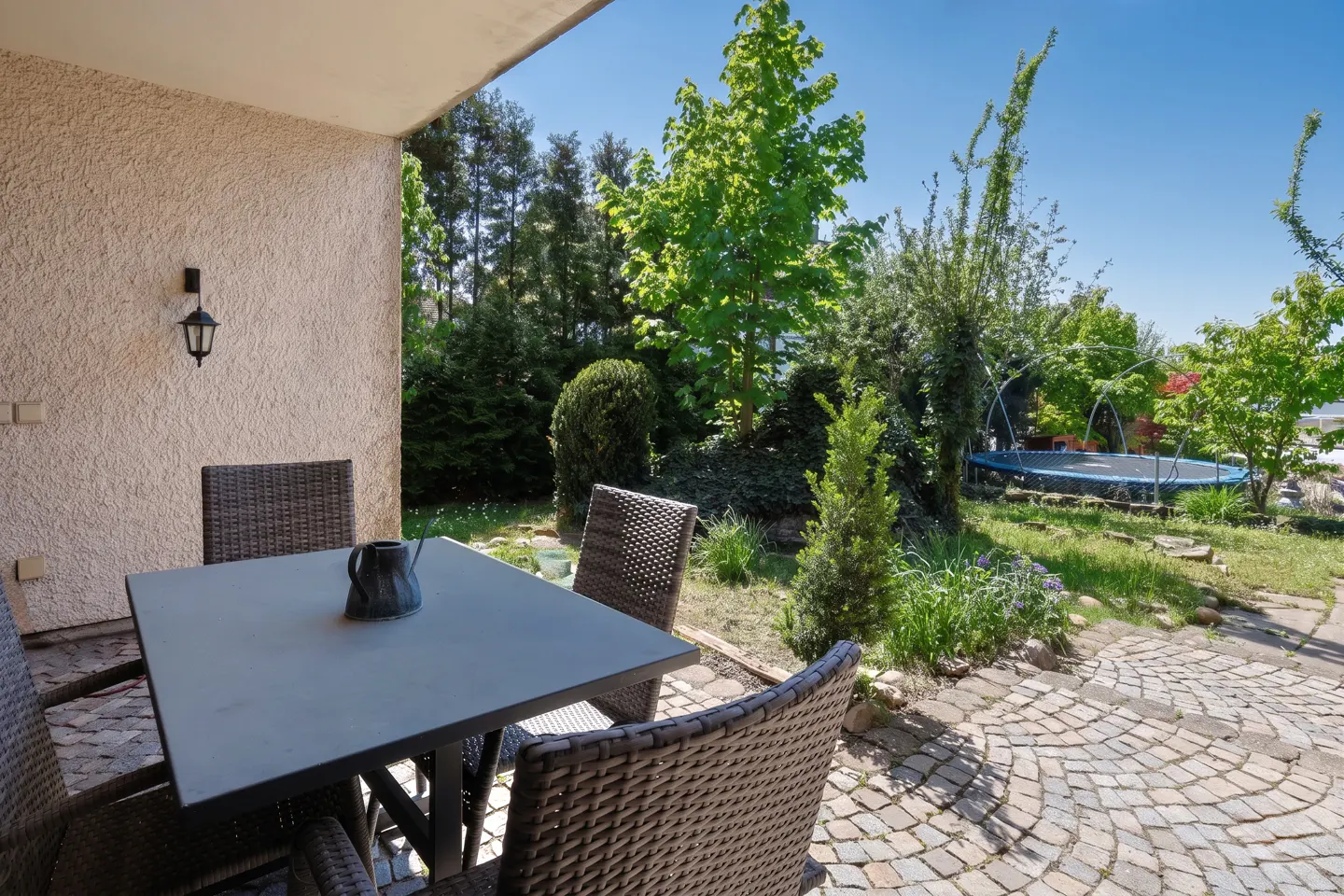 Outdoor patio with a gray table, four wicker chairs, and a black pitcher. A trampoline is visible in the green backyard.