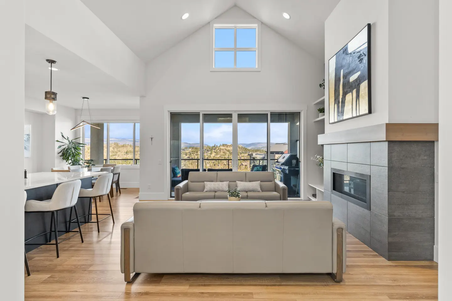 Bright, open-concept living space with hardwood floors, white walls, and vaulted ceiling. Sofas face a fireplace and sliding glass doors to a scenic view.