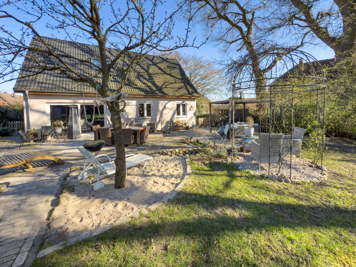 Backyard patio with a beige house, a sand area, and a metal gazebo with chairs on a sunny day.