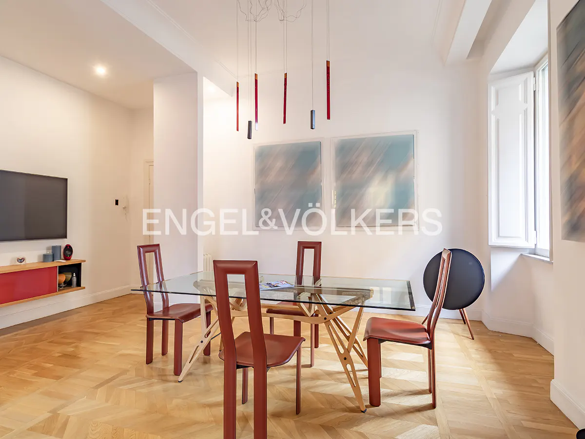 Bright dining room with glass table, red chairs, and modern lighting. Artwork hangs on the white walls. Hardwood floors add warmth.