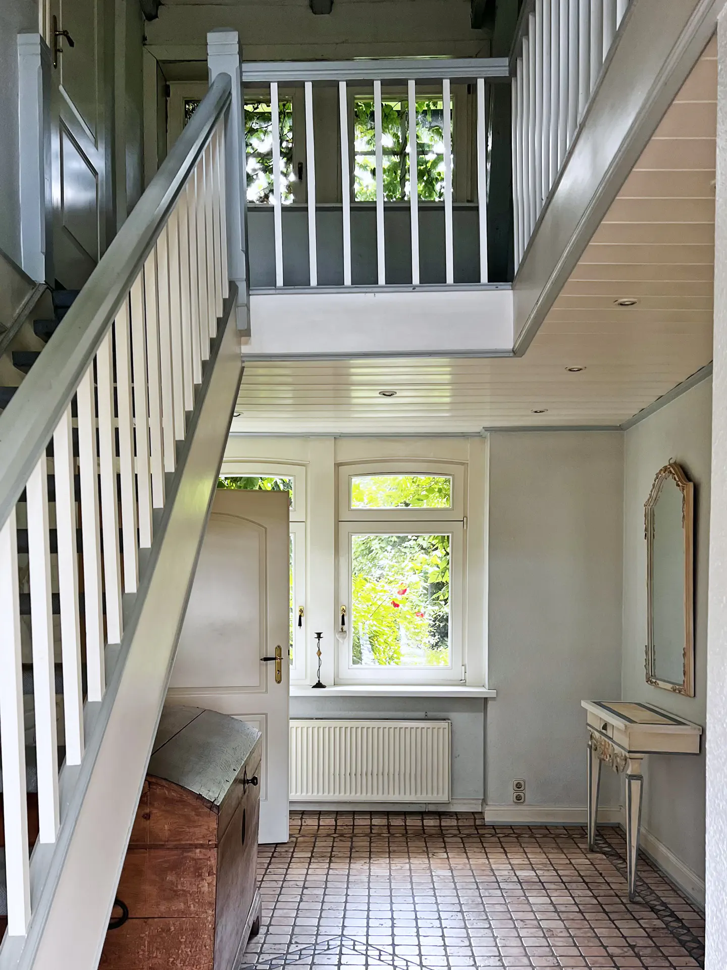 Bright entryway with a wooden staircase, tiled floor, white walls, a console table with a mirror, and garden views.