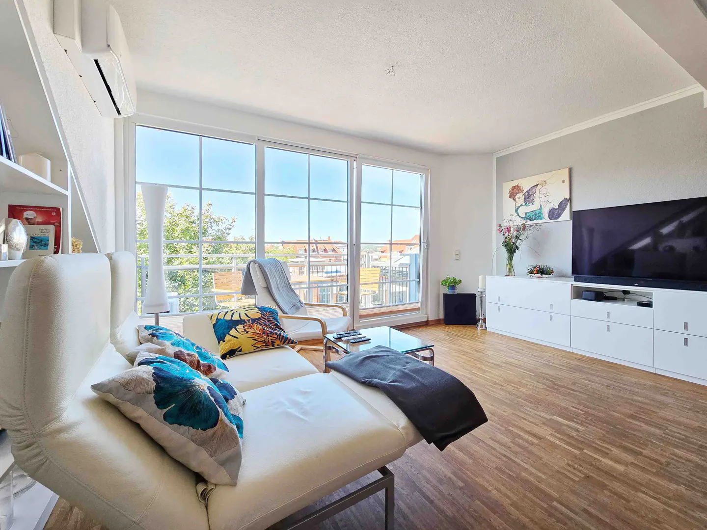 Bright living room with a white sofa, blue pillows, and a large window showing a cityscape. A TV and white cabinet are on the right.