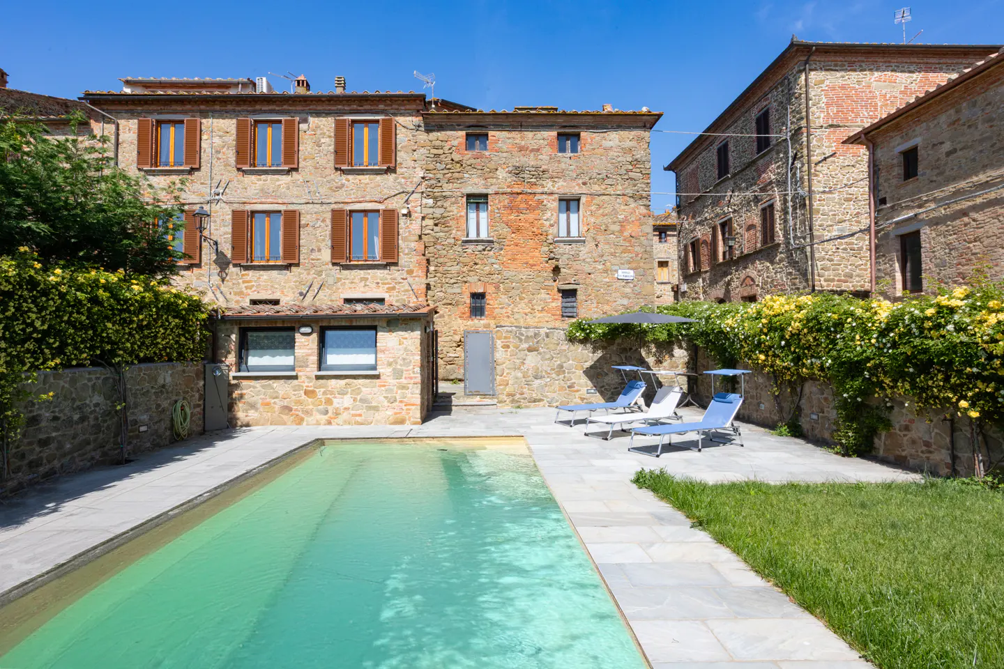 Exterior view of a stone building with a pool, lounge chairs, and green foliage.