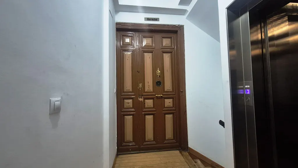 Hallway with a brown wooden door, a metal elevator, and white walls.