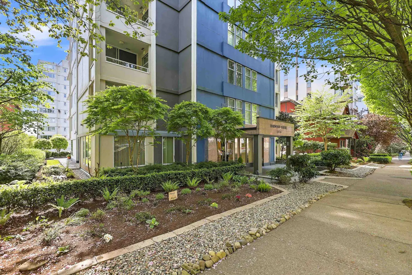 Exterior view of the Harwood House, a blue and beige apartment building with a garden and sidewalk.