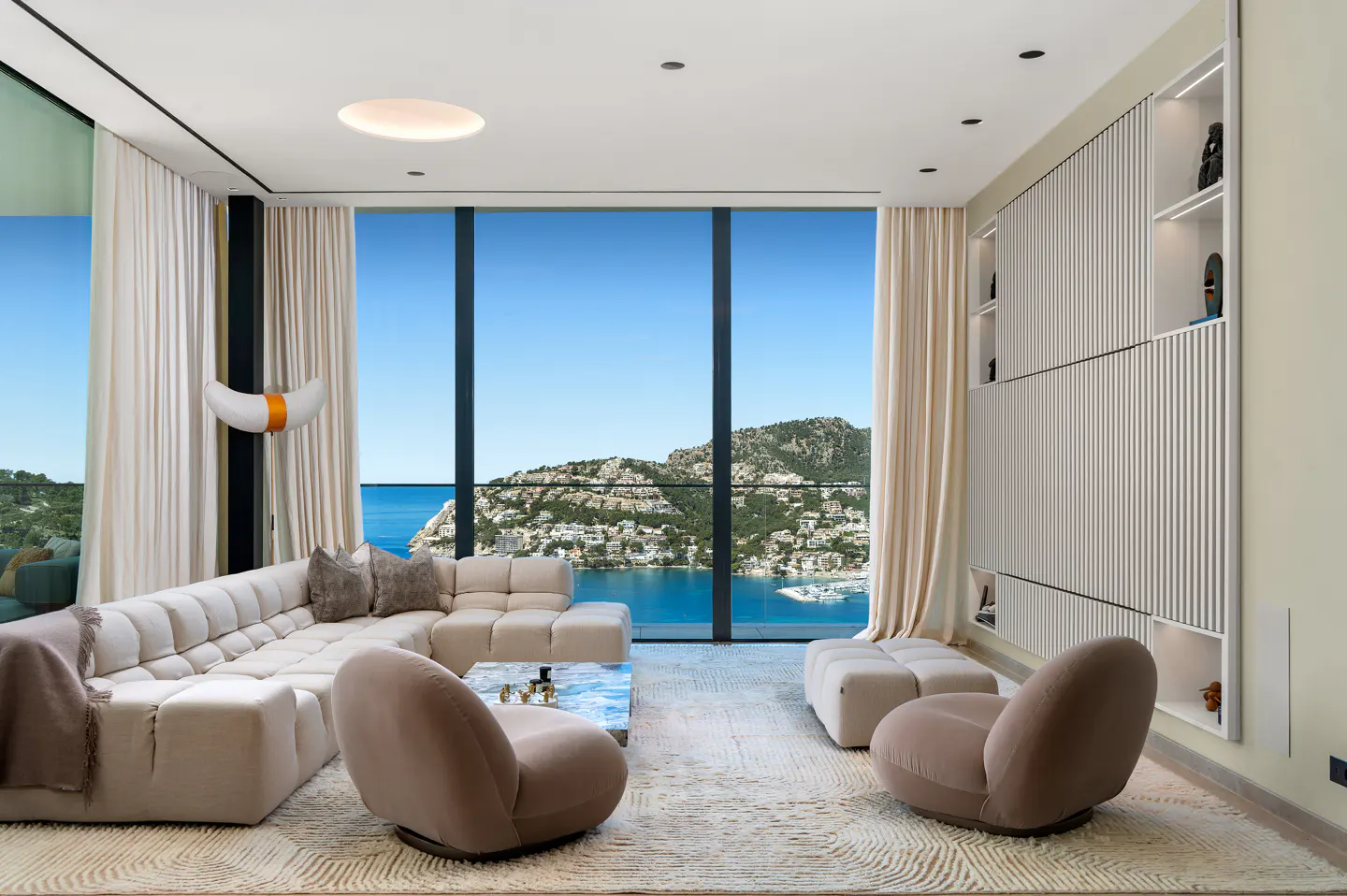 Bright living room with a beige sectional sofa, two chairs, and a large window overlooking the ocean and a coastal town.