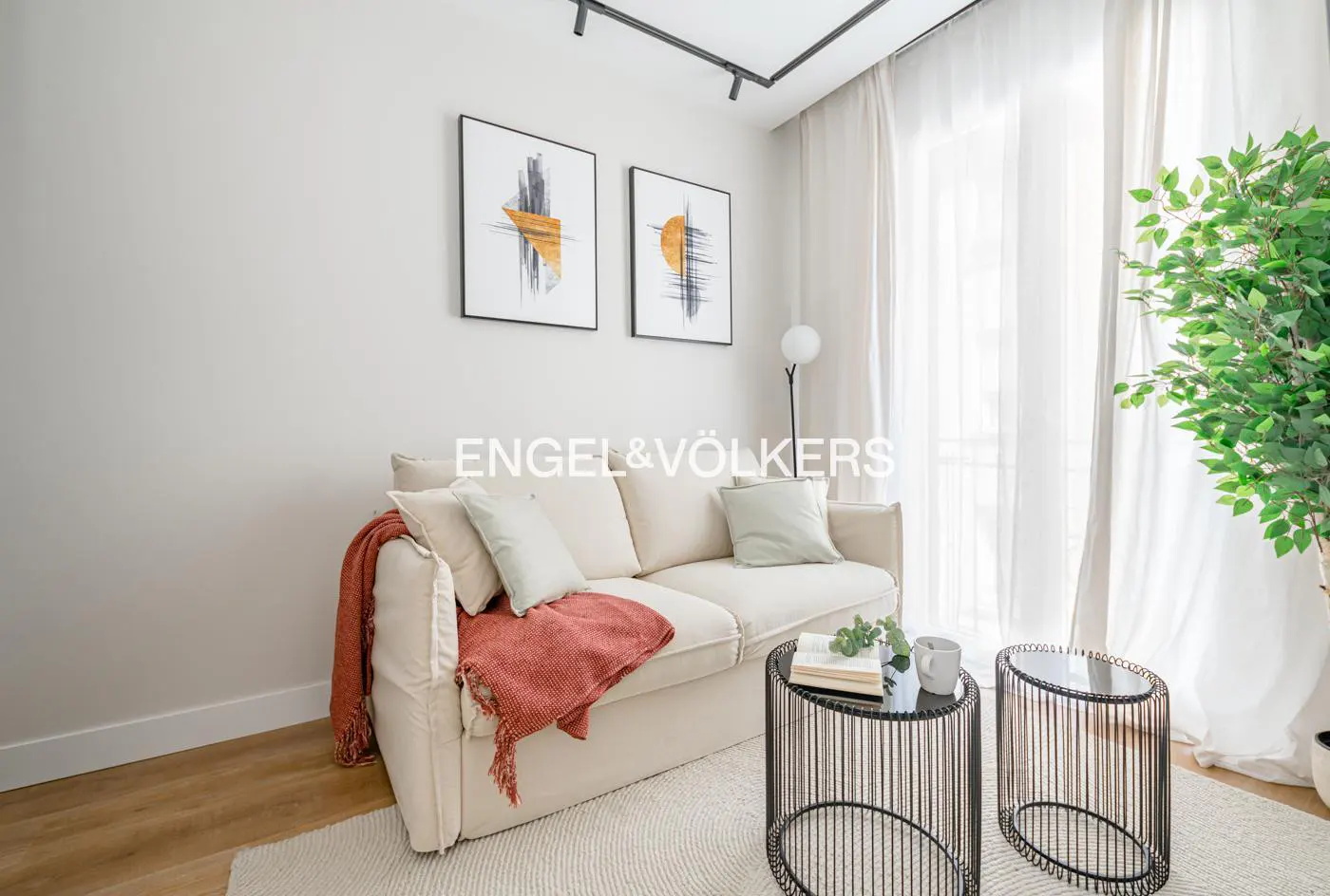 Living room with a cream sofa, rust-colored throw, and two black wire side tables on a white rug. Two framed art prints hang above the sofa.