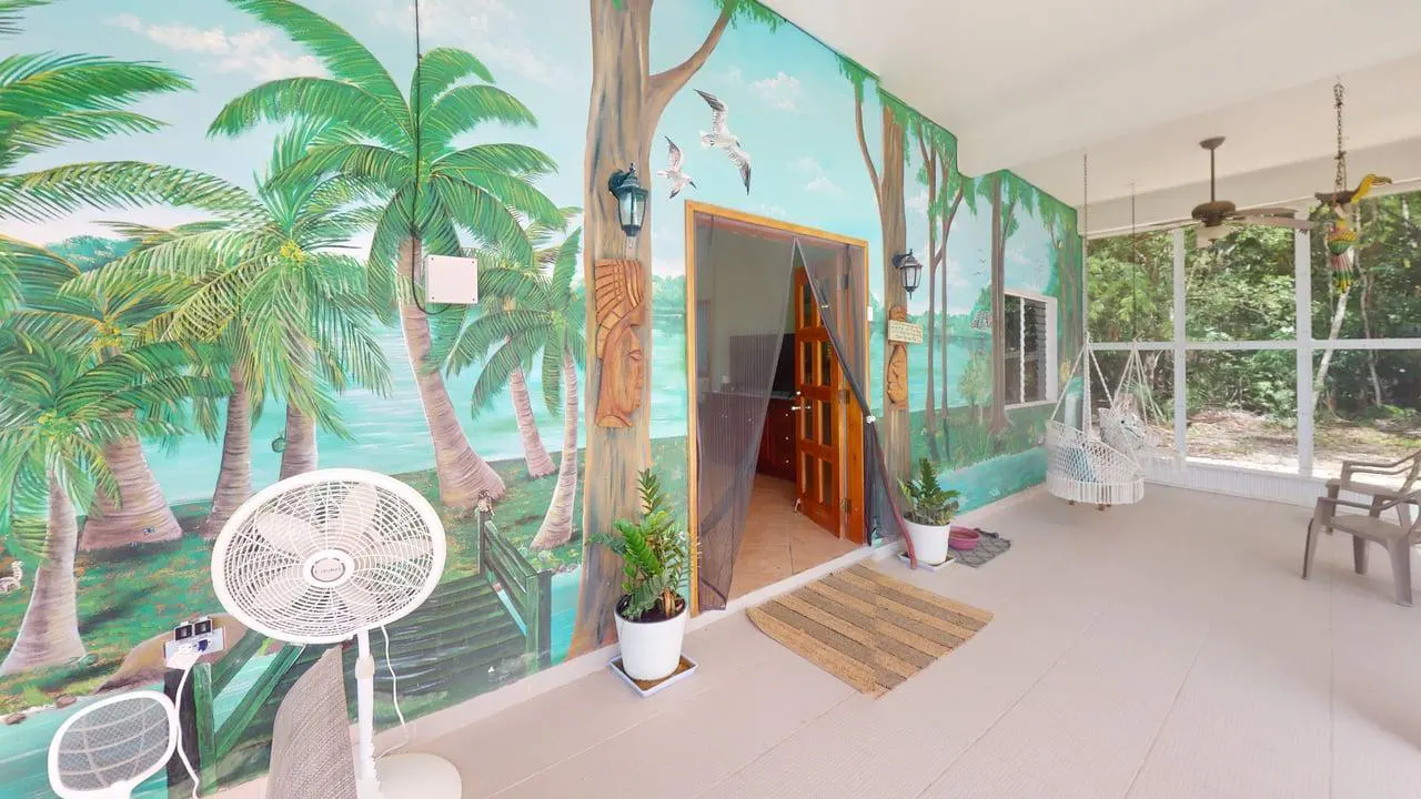 Covered patio with a tropical palm tree mural, a hanging chair, a standing fan, and a doorway leading into the home.