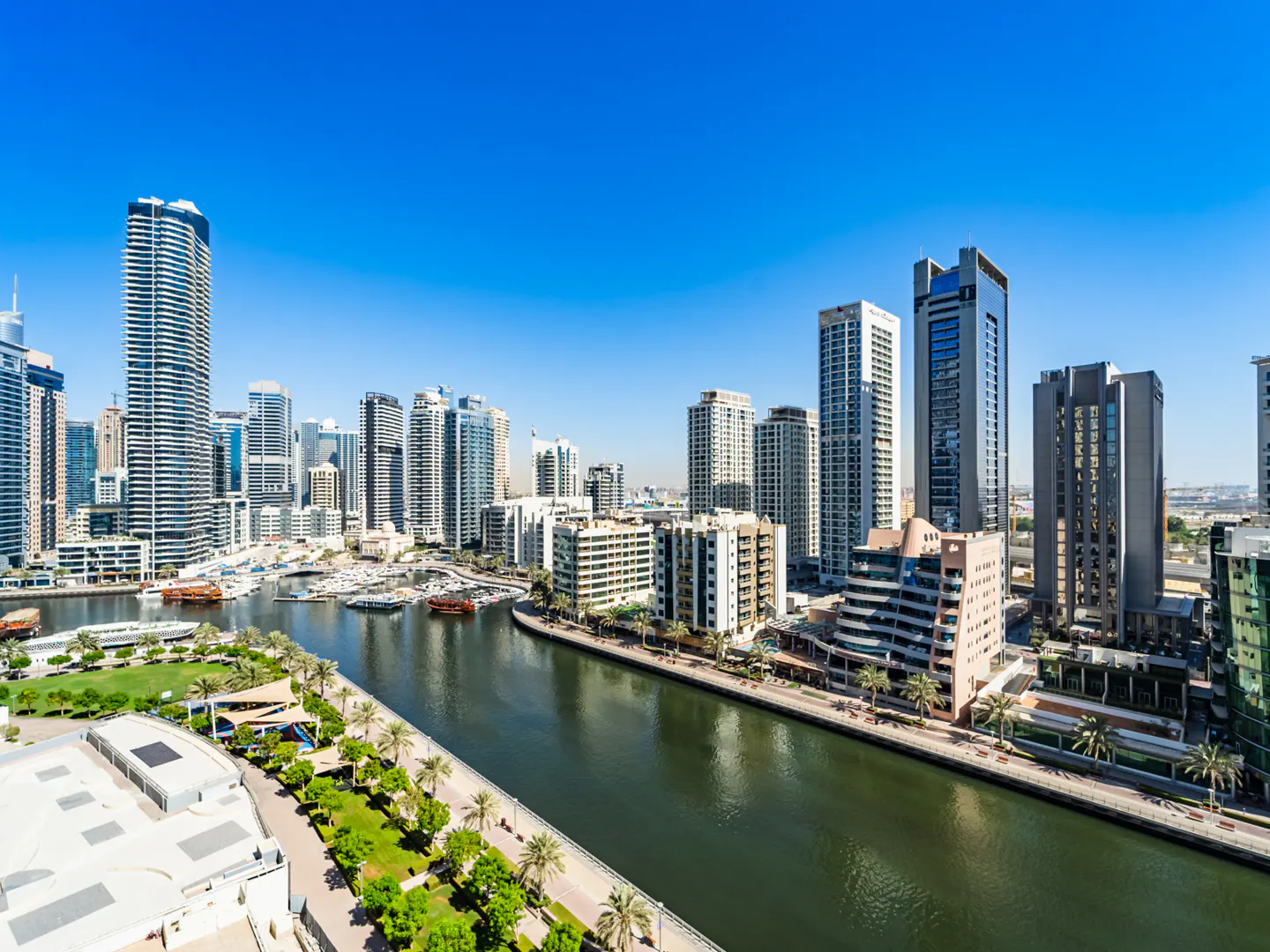 High-angle view of the Dubai Marina canal with modern skyscrapers, palm-lined walkways, and boats under a clear blue sky.