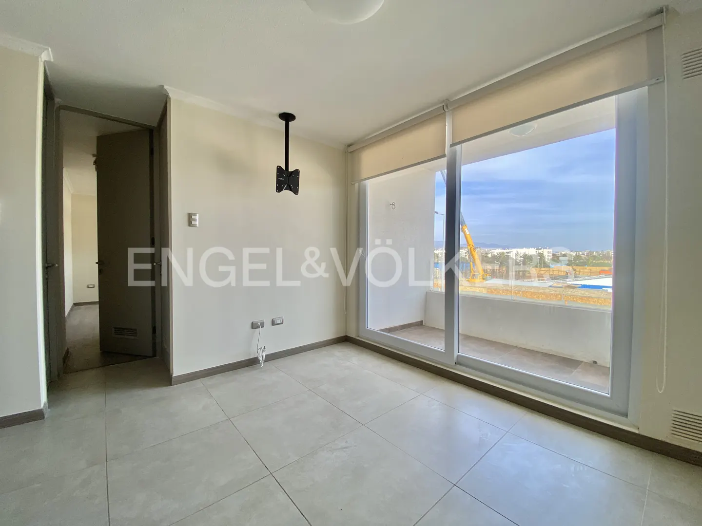 A bright, empty room with gray tile floors, white walls, and a large window with a view of a city and construction. A door leads to another room.