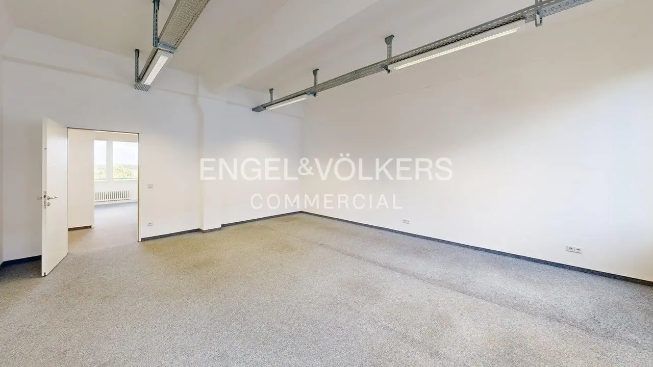 Empty commercial space with gray carpet, white walls, and fluorescent lighting. An open doorway leads to another room. "Engel & Völkers Commercial" logo on the wall.