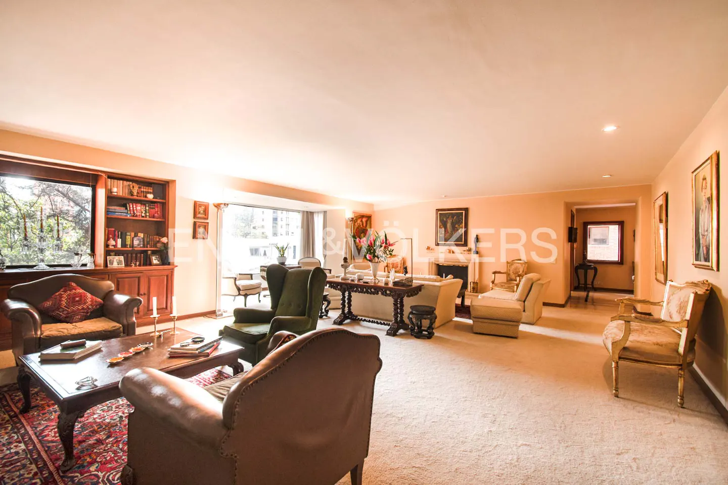 Spacious living room with beige carpet, brown leather chairs, and a dark wood coffee table on a red patterned rug. A bookcase and large window are on the left.