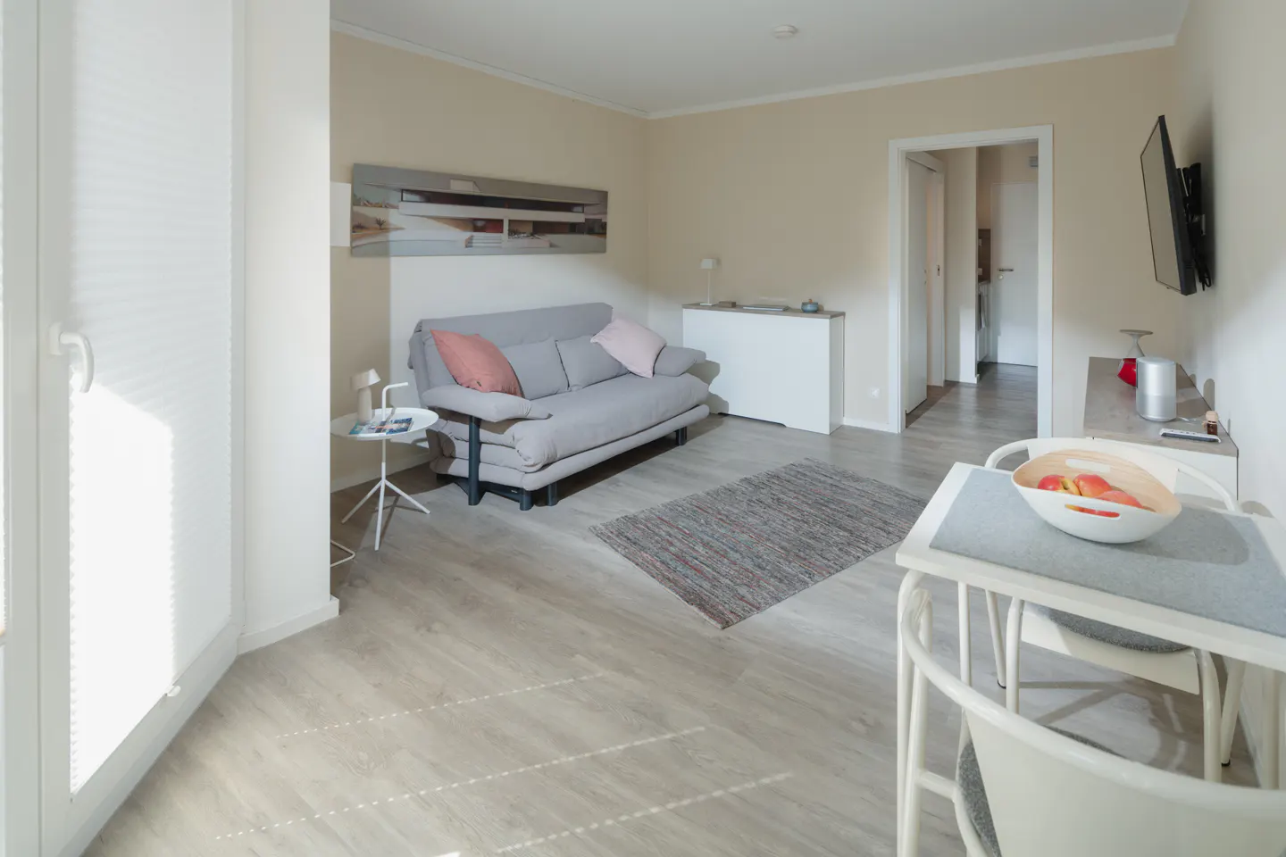 A bright living room with a gray sofa, a small table, and a white dining table with a fruit bowl.