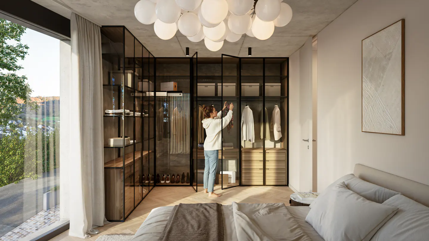 A woman in a white hoodie organizes clothes in a modern walk-in closet with black framed glass doors, next to a bed and a large window.