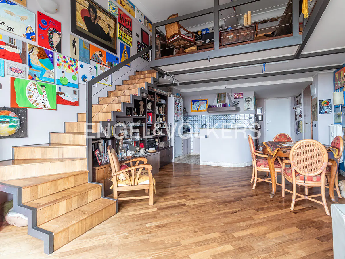 Interior view of a modern apartment with wooden floors, stairs to a loft, and walls covered in colorful artwork. A dining table with chairs is visible.