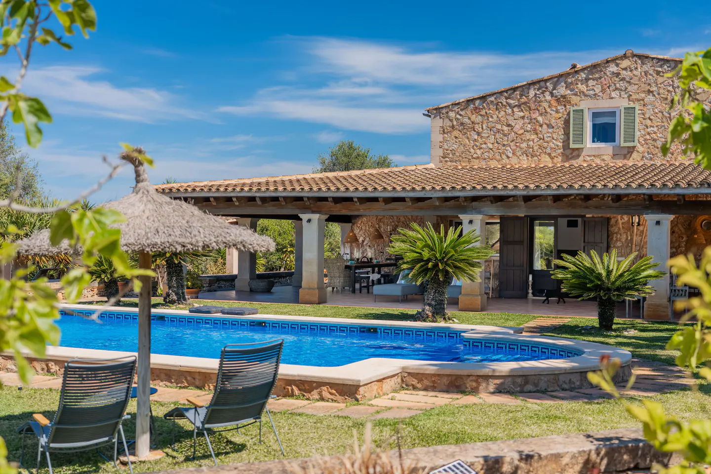 Exterior view of a stone house with a blue pool, two chairs, and a straw umbrella on a sunny day.
