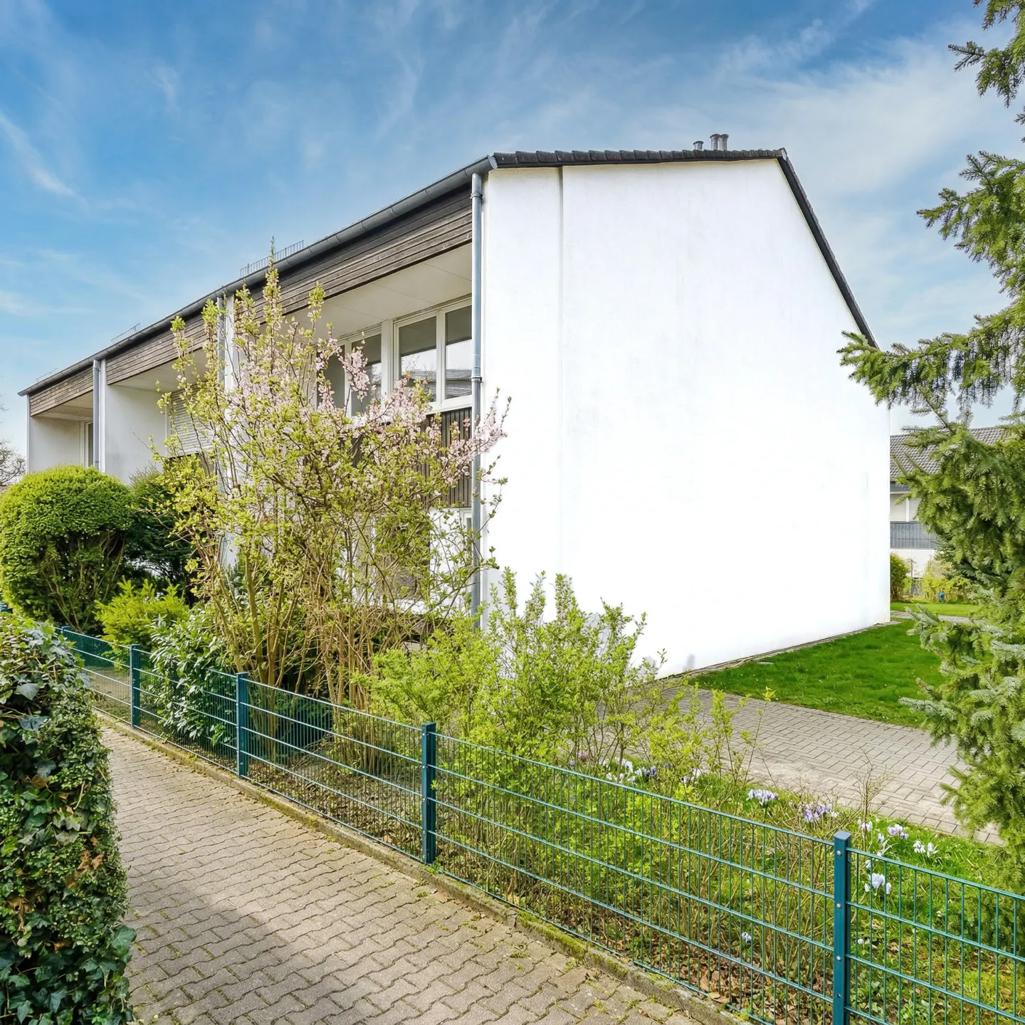 Exterior view of a white, two-story townhouse with a green metal fence and a brick walkway. Bushes and trees surround the property.