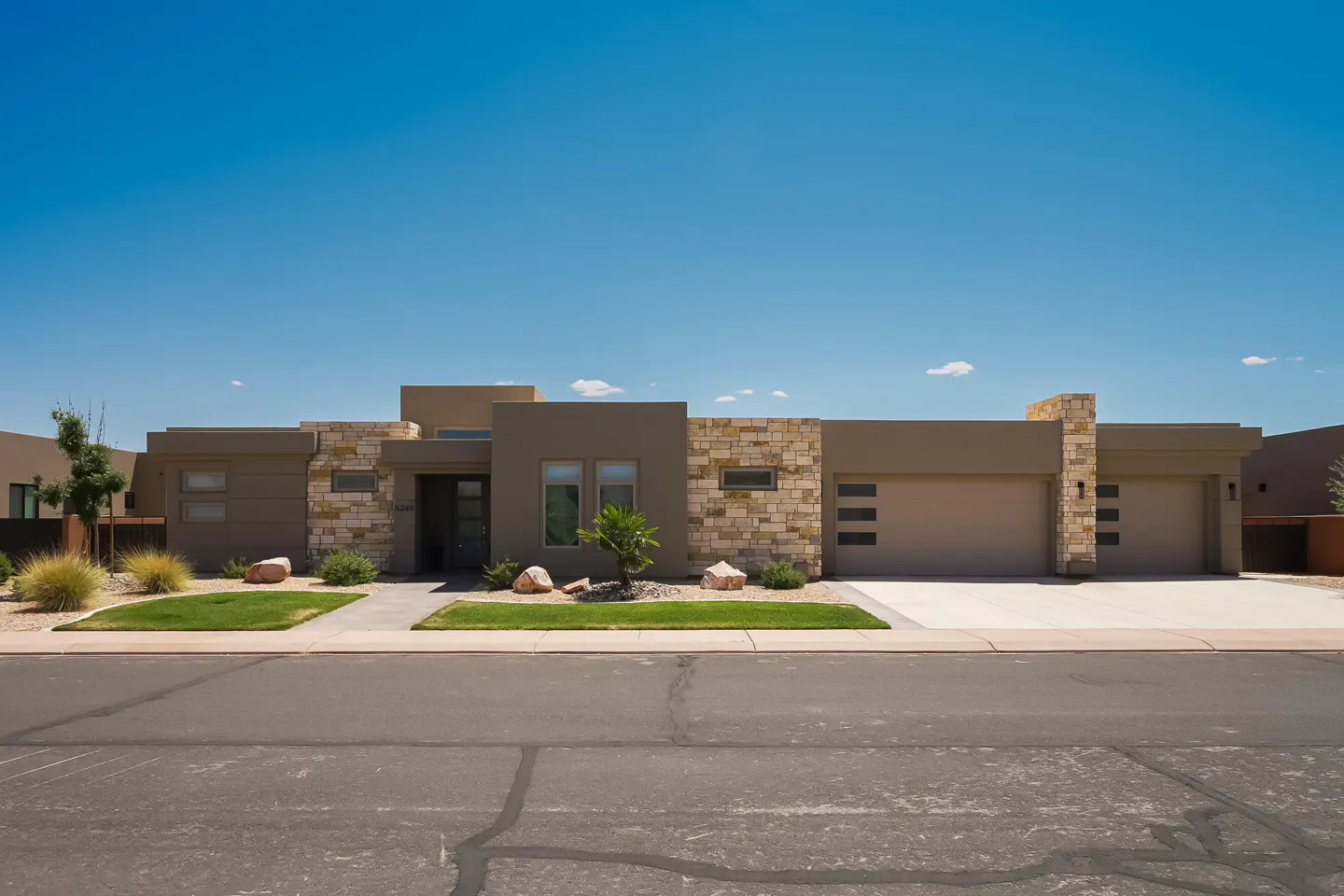 Modern single-story home with a two-car garage, tan stucco, stone accents, and a well-manicured lawn under a clear blue sky.