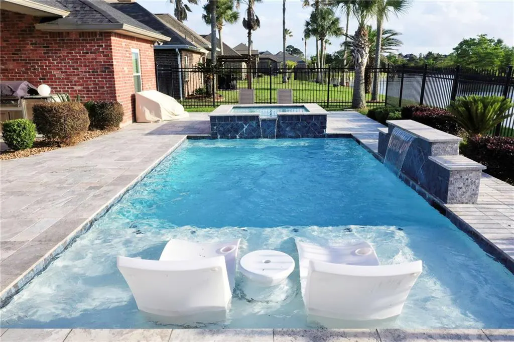 A backyard pool with two white chairs and a table in the water. A waterfall feature and a hot tub are in the background.