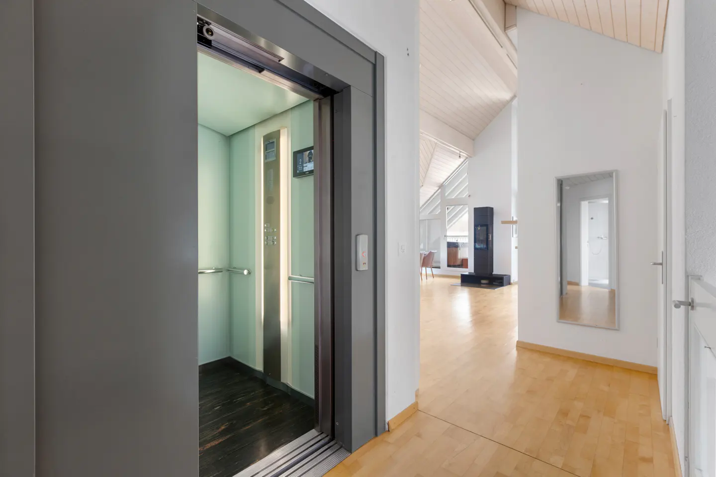 Open elevator with gray frame and light green interior, dark wood floor. Hallway with wood floors, white walls, mirror, and wood-paneled ceiling.