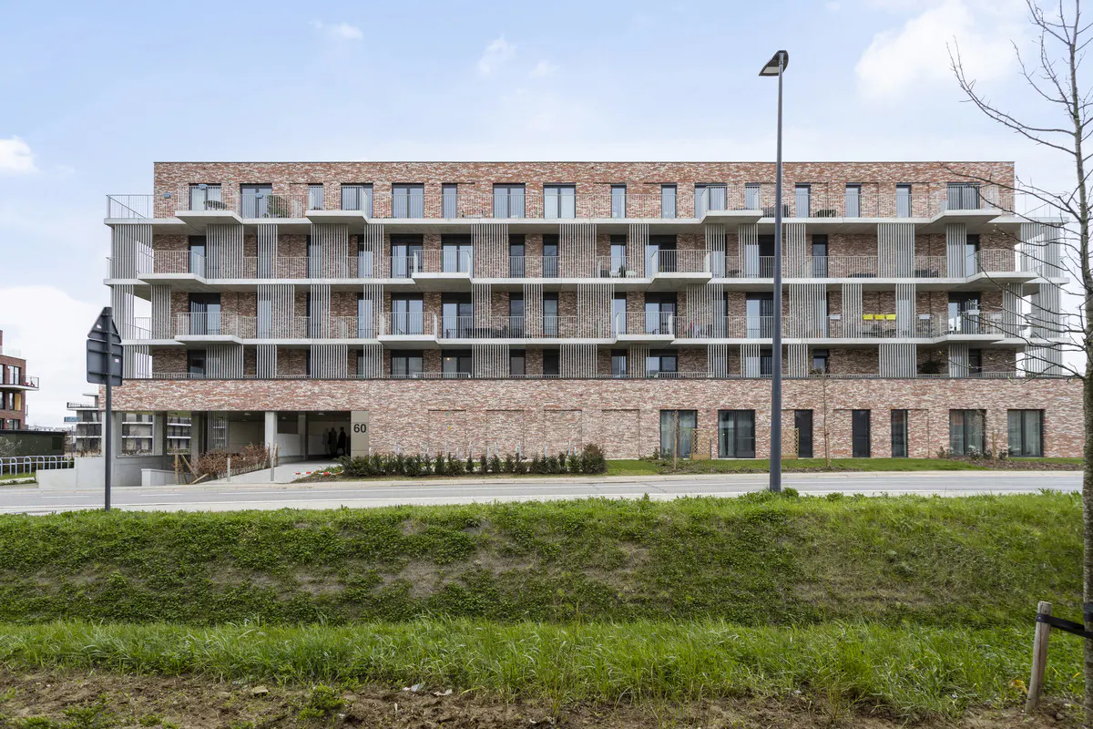 Modern brick apartment building with balconies and large windows, set against a blue sky and green landscape.