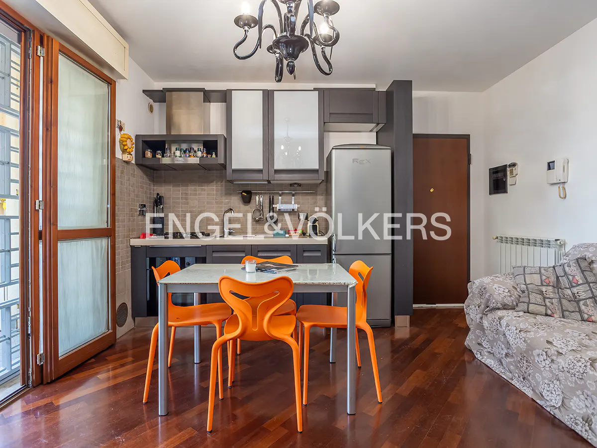 A modern apartment interior with a kitchen, dining table with orange chairs, and a floral sofa.