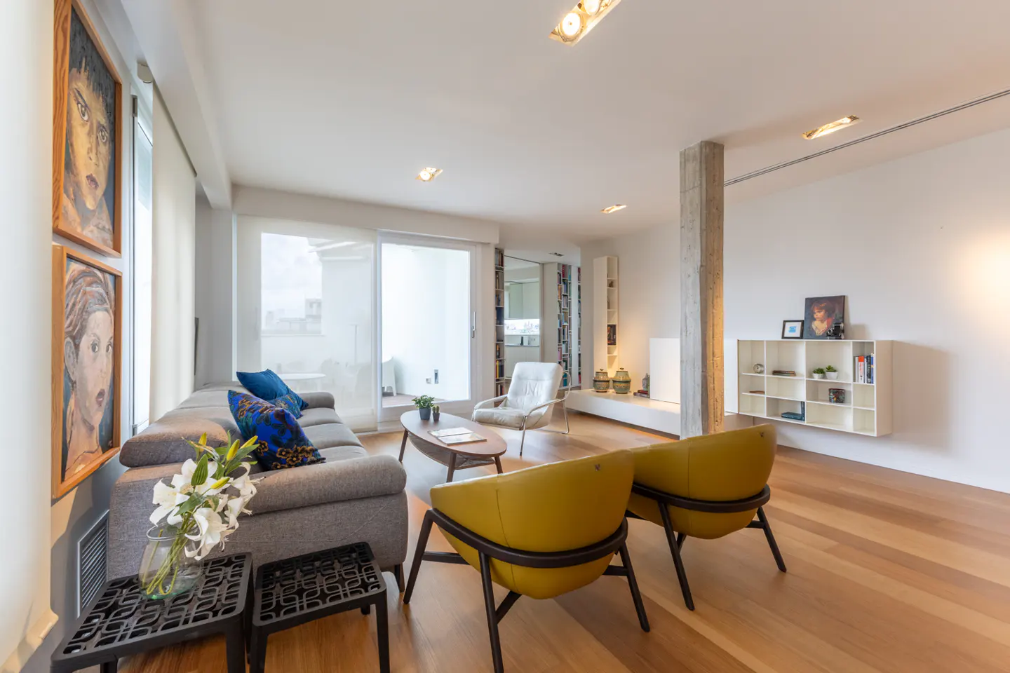 Bright living room with wood floors, gray sofa with blue pillows, and two yellow chairs. Artwork on the wall and a balcony in the background.