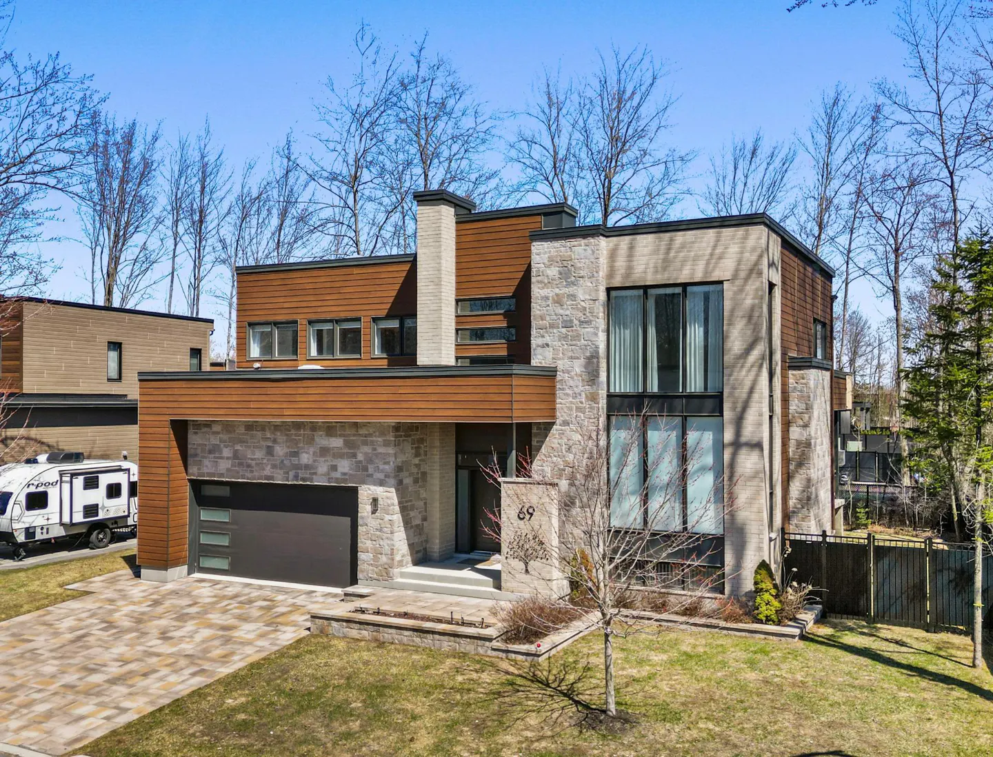 Modern two-story home with stone and wood facade, a dark garage door, and a paved driveway. A small camper is parked nearby.