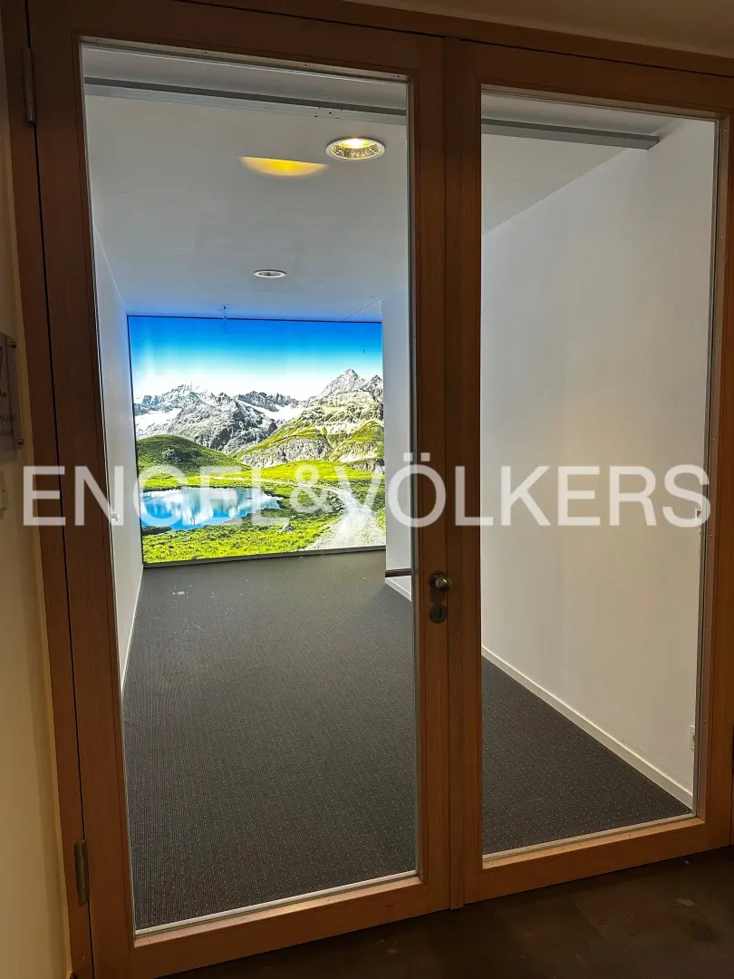 Interior view through glass doors into two rooms with gray carpet. One room has a mountain mural. Engel & Völkers logo is visible.