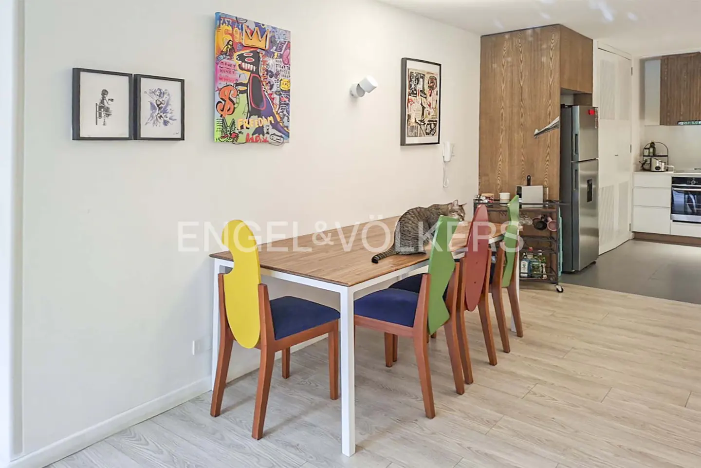 A dining room with a cat on the table. The chairs are colorful and the walls have art.