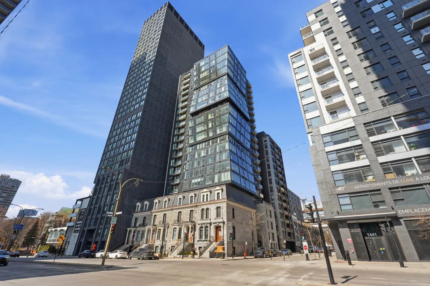 Street-level view of modern buildings in Montreal, Canada, under a blue sky. A tall, dark skyscraper stands next to a glass-fronted building.