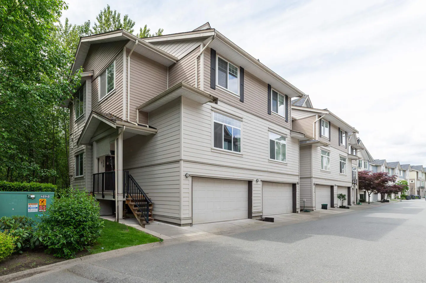 Exterior view of a multi-story beige townhouse complex with attached garages and a small staircase, set along a paved road.