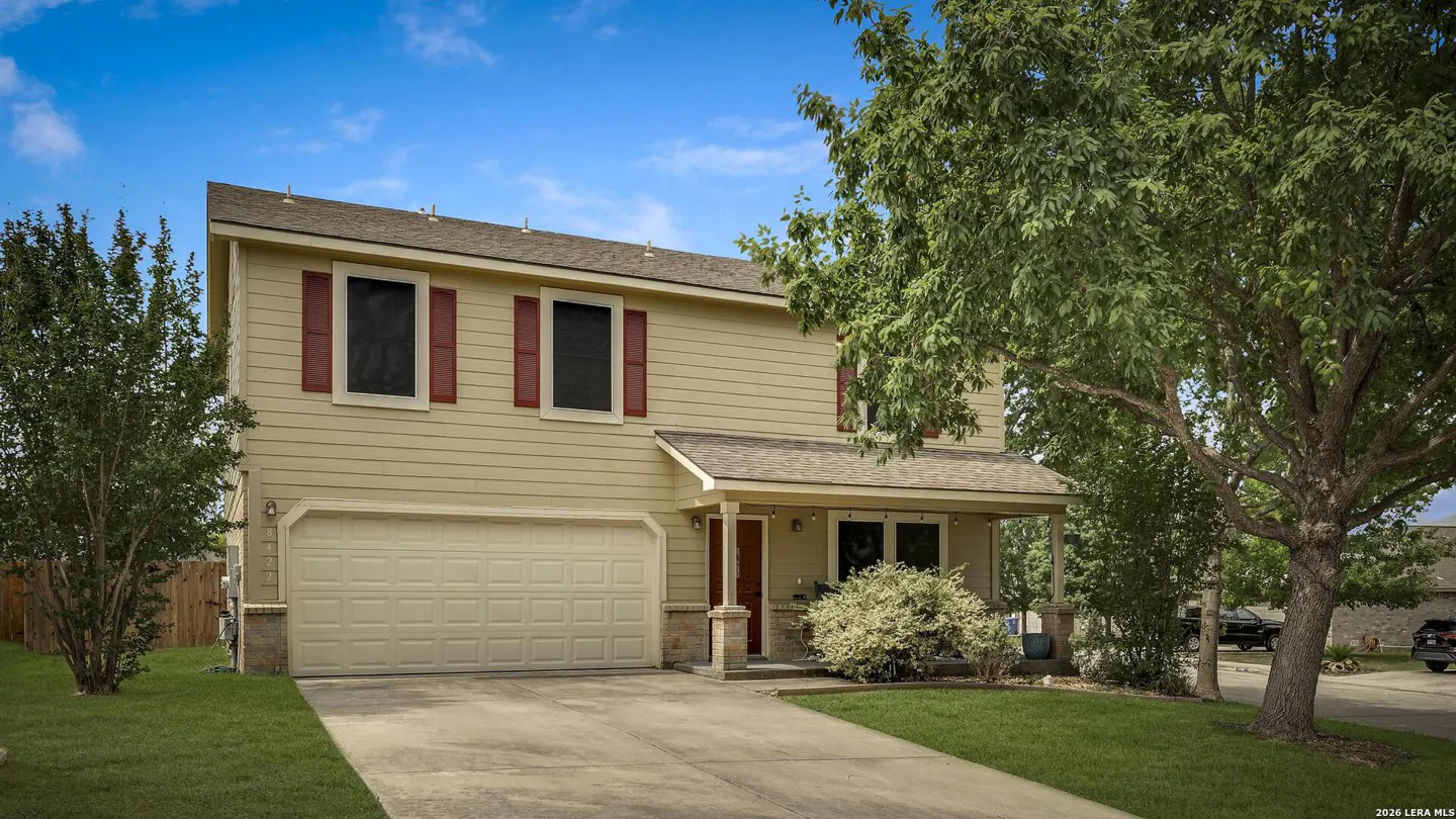 Two-story beige house with red shutters, a two-car garage, and a green lawn under a blue sky.