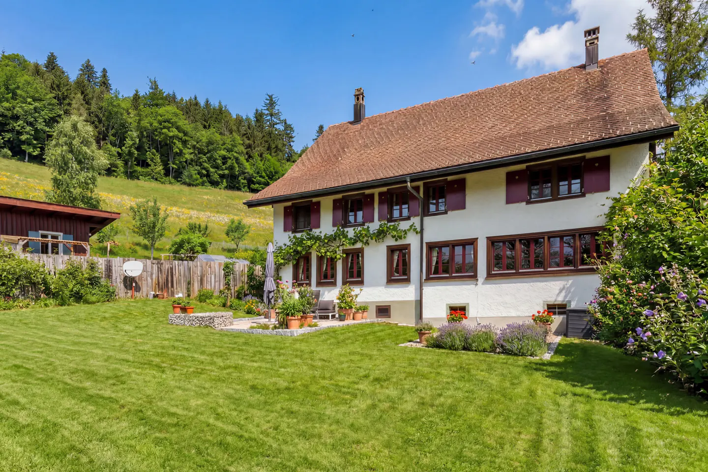 White two-story house with brown shutters and a tiled roof, set in a lush green garden against a forested hillside.