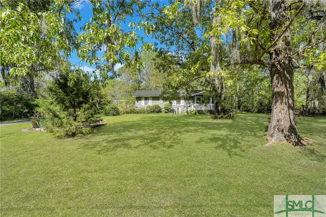 A quaint, light-gray house with a white porch is nestled among trees with Spanish moss, on a large green lawn.