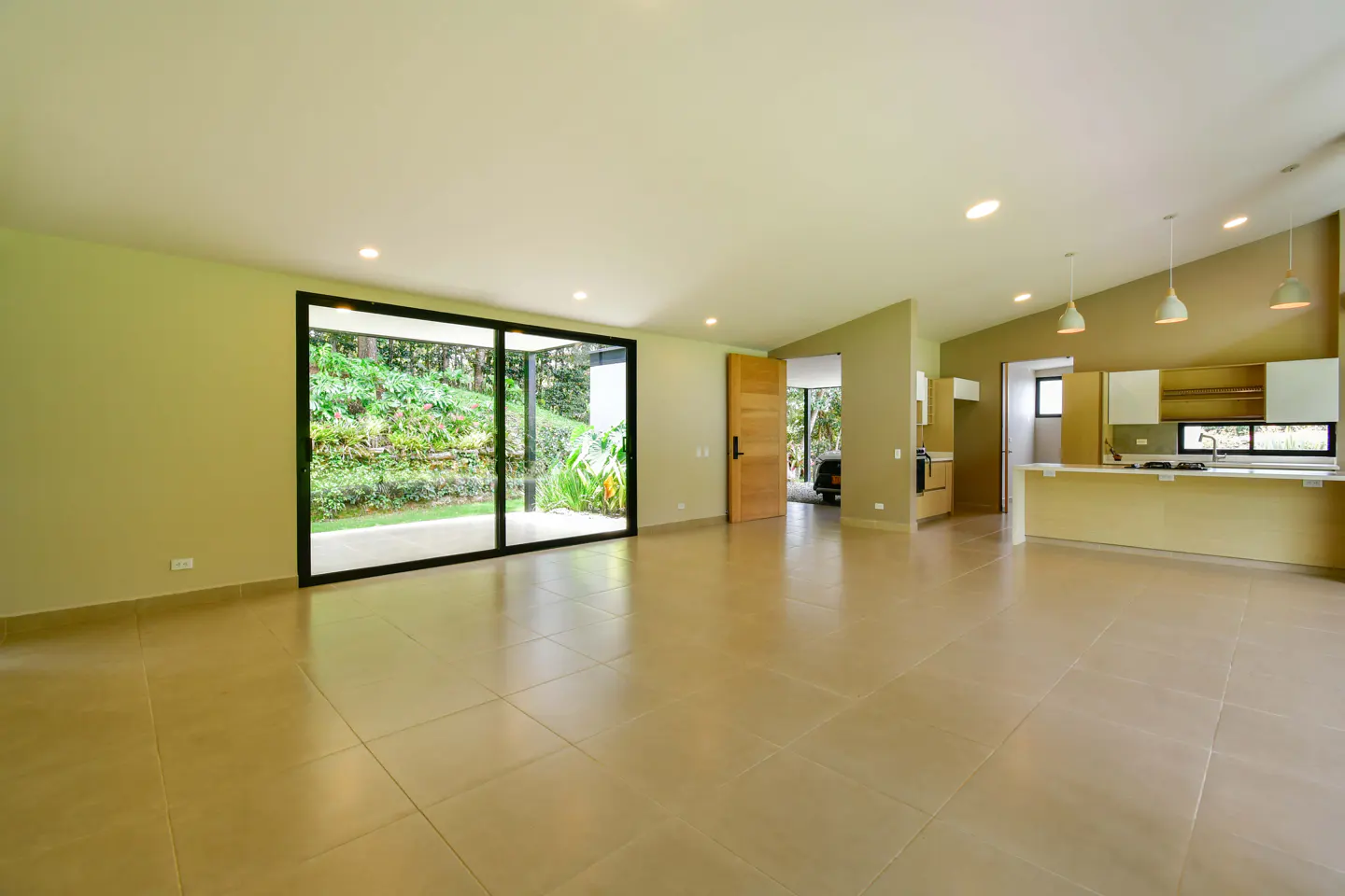 Open-concept living space with beige tile flooring, a kitchen with white cabinets, and a large sliding glass door to a lush green yard.