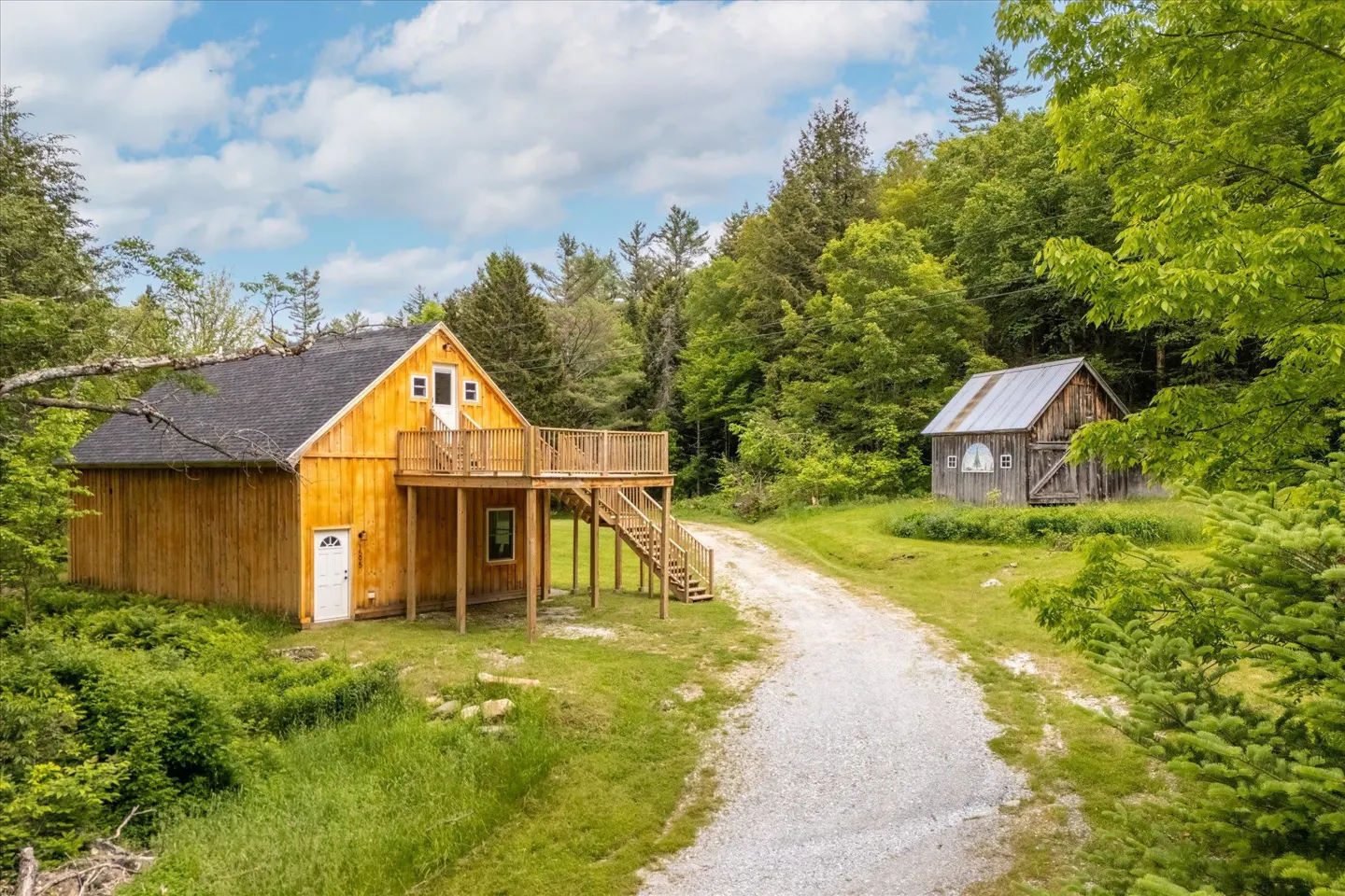 Wooden house with a deck and stairs, surrounded by green trees and grass, with a gravel driveway leading to a small barn.