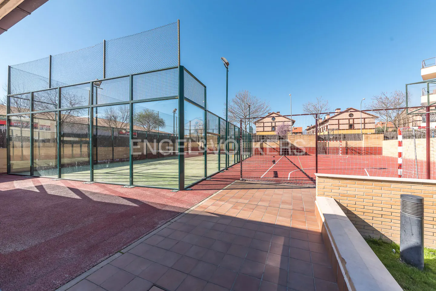 Outdoor view of a padel court with green glass walls and a red floor, next to a red basketball court.