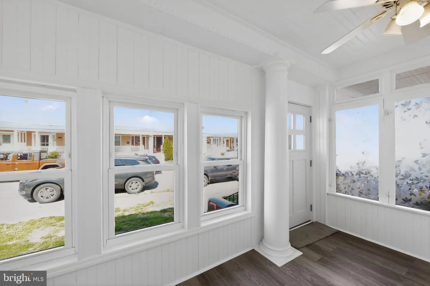 Bright sunroom with white walls, dark wood floors, and large windows. Cars and buildings are visible outside. A white column and door are on the right.