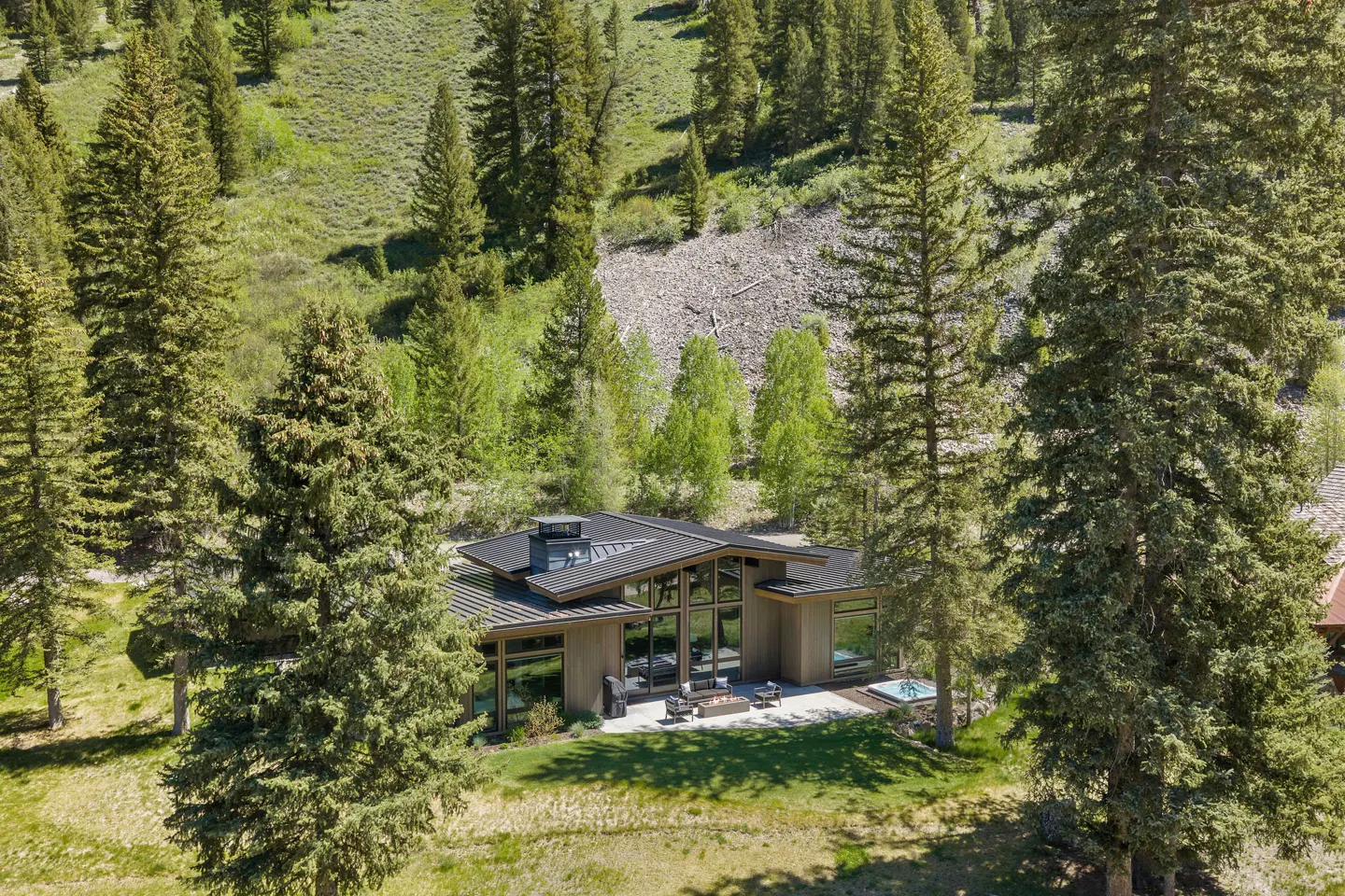 Modern home with a black roof and large windows, nestled among tall green trees on a grassy hillside.