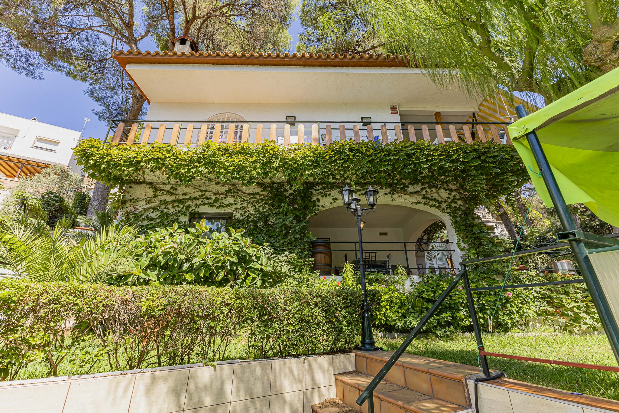 Two-story white house covered in ivy with a balcony, arched patio, and tiled stairs in a lush, sunny garden setting. [AI-generated text]