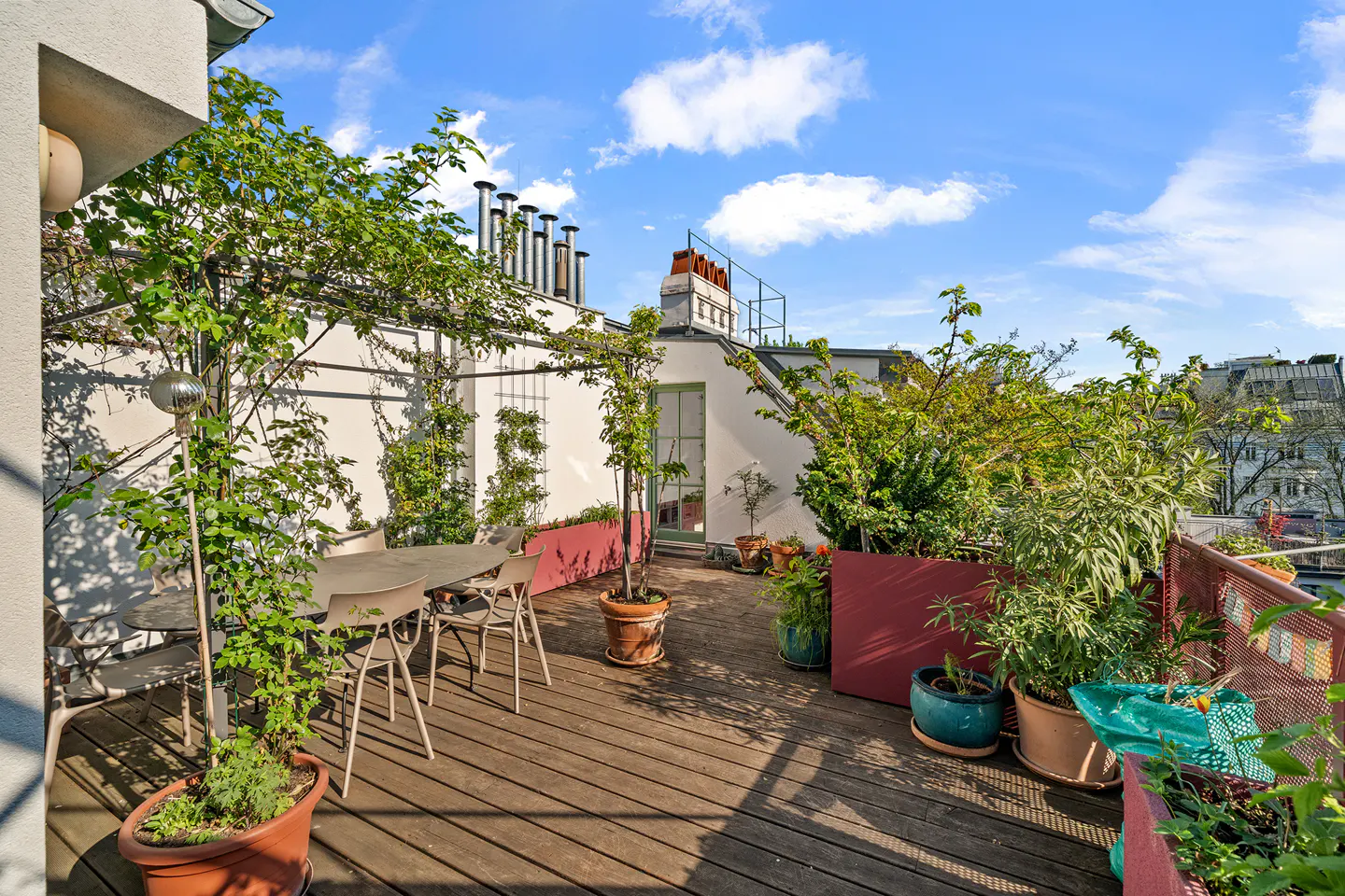 Rooftop patio with wooden deck, table, chairs, plants in pots, and white walls under a blue sky.