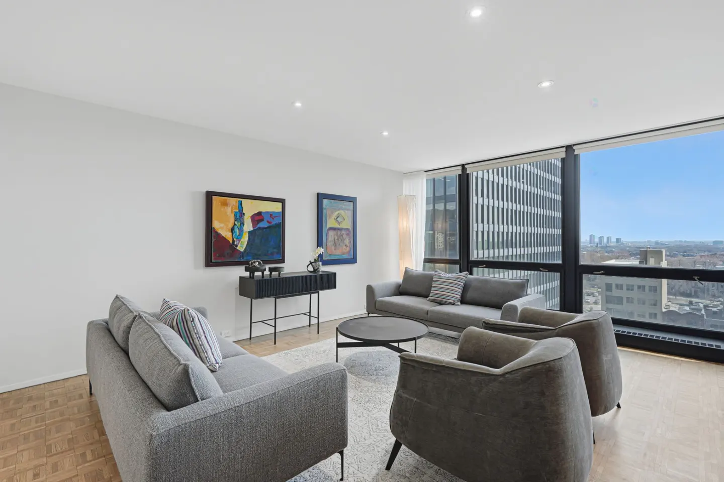 Bright living room with gray sofas, chairs, and a black console table with colorful art. Large windows show a city view.