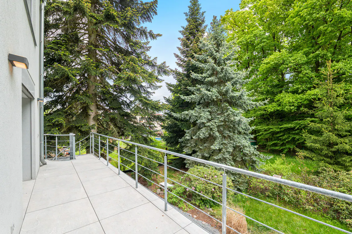 Balcony view with metal railing, light gray tile floor, and lush green trees in the background under a blue sky.