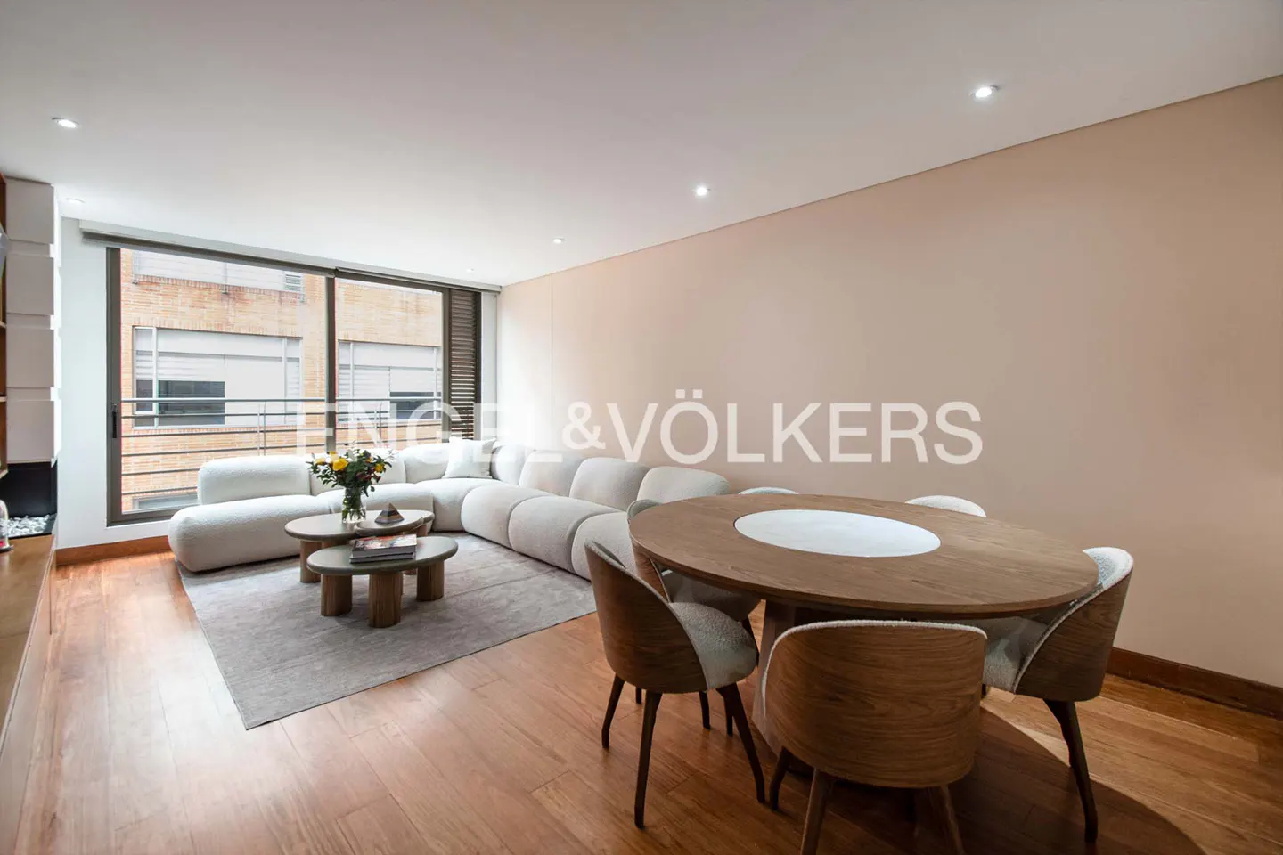 Modern living room with a white sectional sofa, wooden dining table, and large windows overlooking a city building.